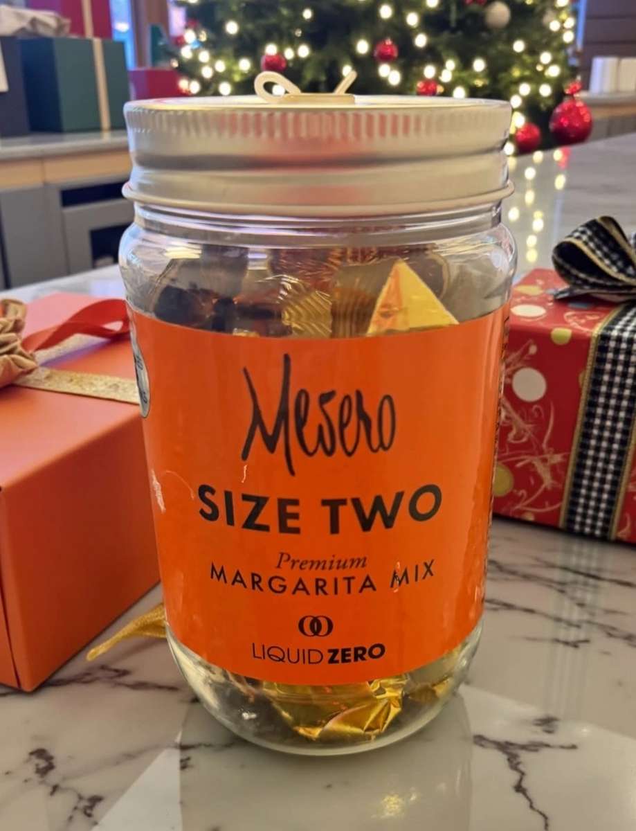 Mesero size two premium Margarita mix jar with dried citrus inside, set against festive holiday lights and wrapped gifts.