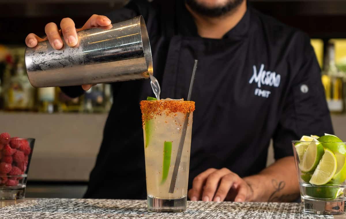 A Mesero bartender pouring a cocktail with a chili rim and lime garnish at the bar counter.