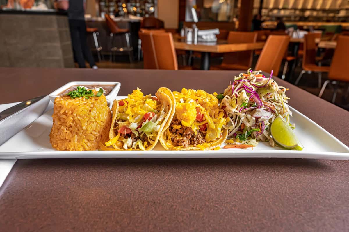 Three beef tacos with shredded cheese and lettuce, served with seasoned rice, on a rectangular white plate at Mesero.