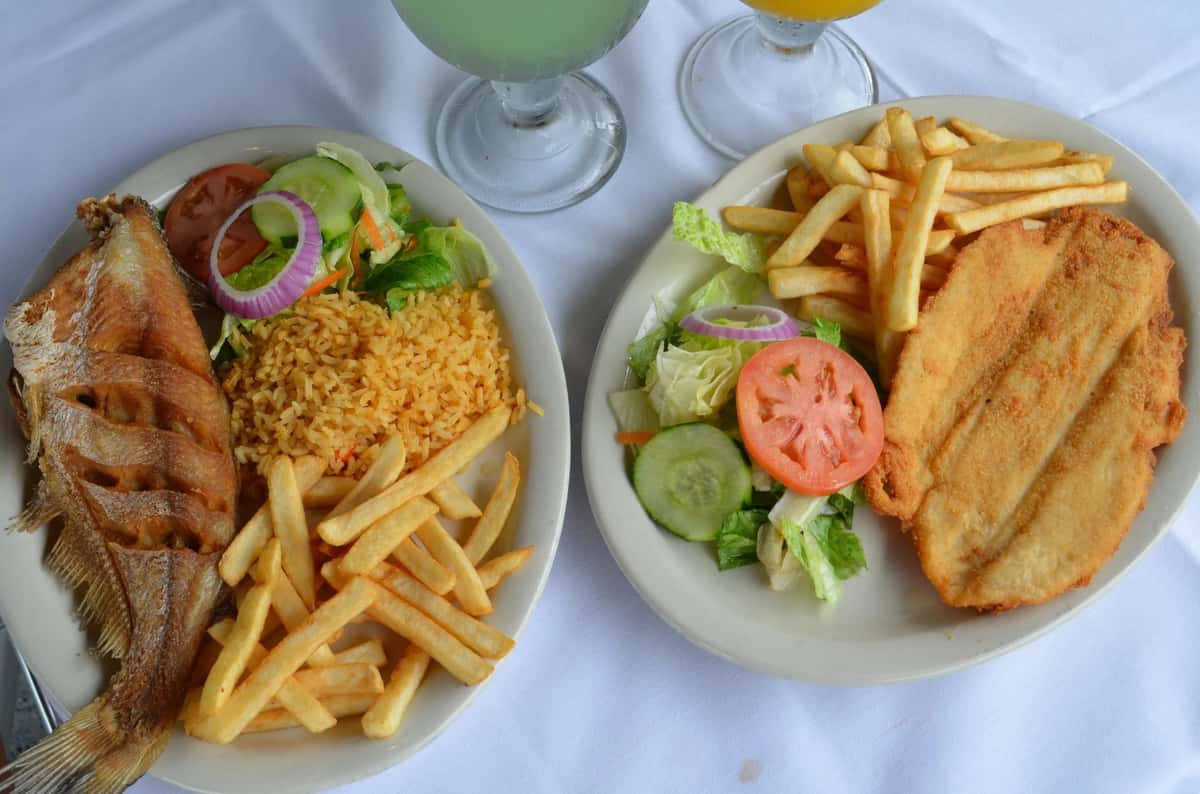 Two Mexican seafood platters—one with whole fried fish and the other with breaded fish fillet—served with golden fries, fresh garden salad, Mexican rice, and refreshing aguas frescas on a white table.