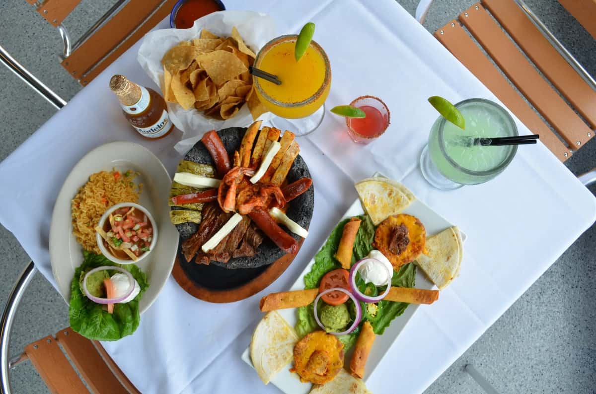 Vibrant Mexican feast featuring a sizzling molcajete with grilled meats and shrimp, crispy taquitos, quesadillas, pico de gallo, tortilla chips, and colorful margaritas on a white tablecloth patio setting.