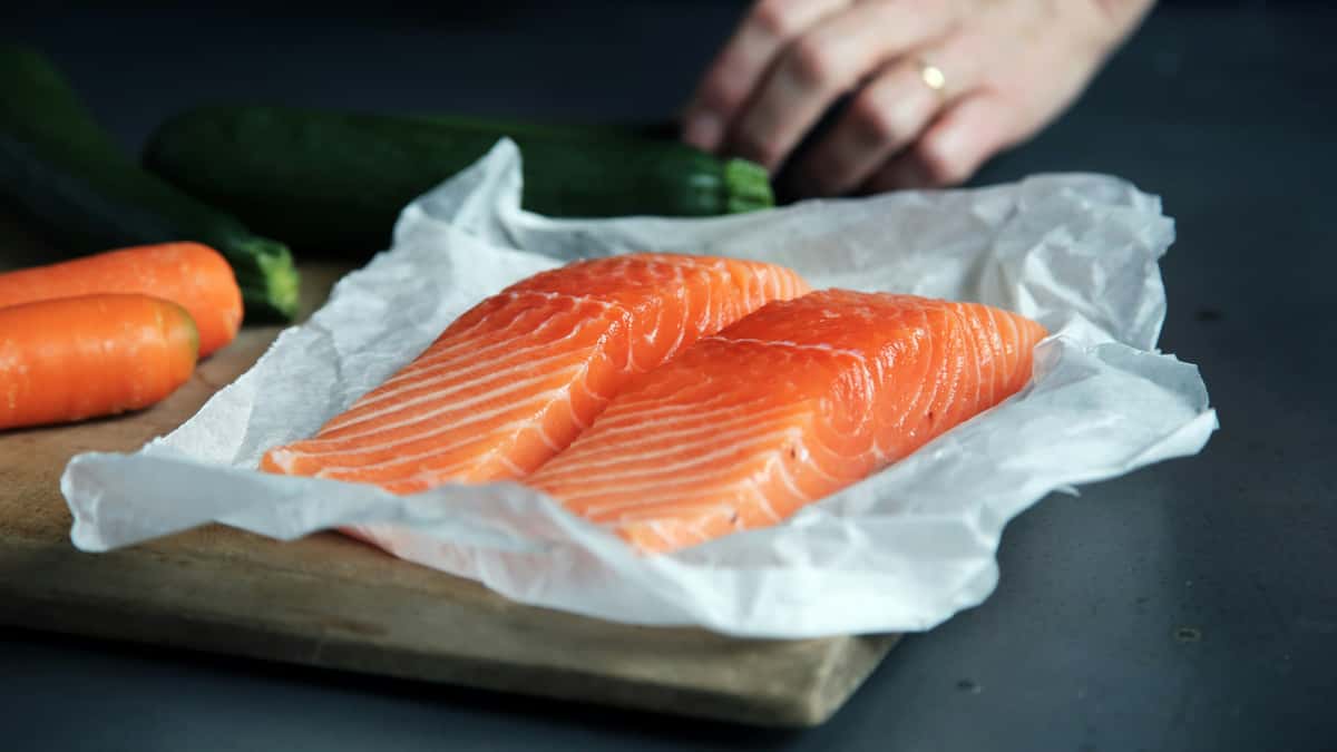 Two fresh, raw salmon fillets with rich orange marbling placed on parchment paper, with carrots and zucchini in the background—ideal for sushi, grilling, or seafood recipes.