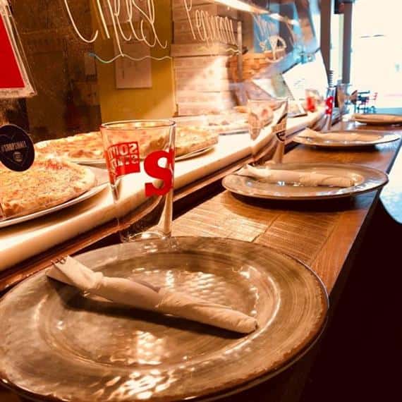 Plate on a counter-top with a fork and knife wrapped in a napkin and an empty beer glass with assorted pizzas behind a glass display