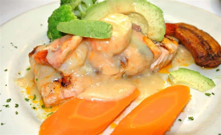 Grilled salmon topped with shrimp, avocado, and a creamy sauce with broccoli and sliced carrots on the side