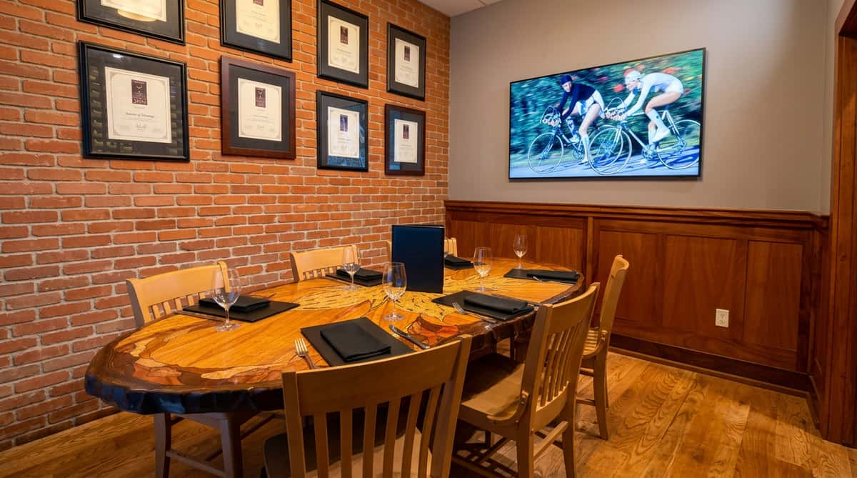 Conference room with a large wooden table set for eight, brick wall with framed awards, and a wall-mounted TV showing cyclists.