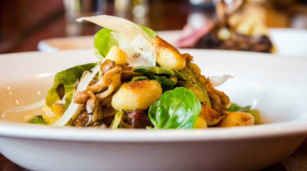 Gnocchi with sautéed mushrooms, green leafy vegetables, and shaved parmesan cheese in a white bowl.