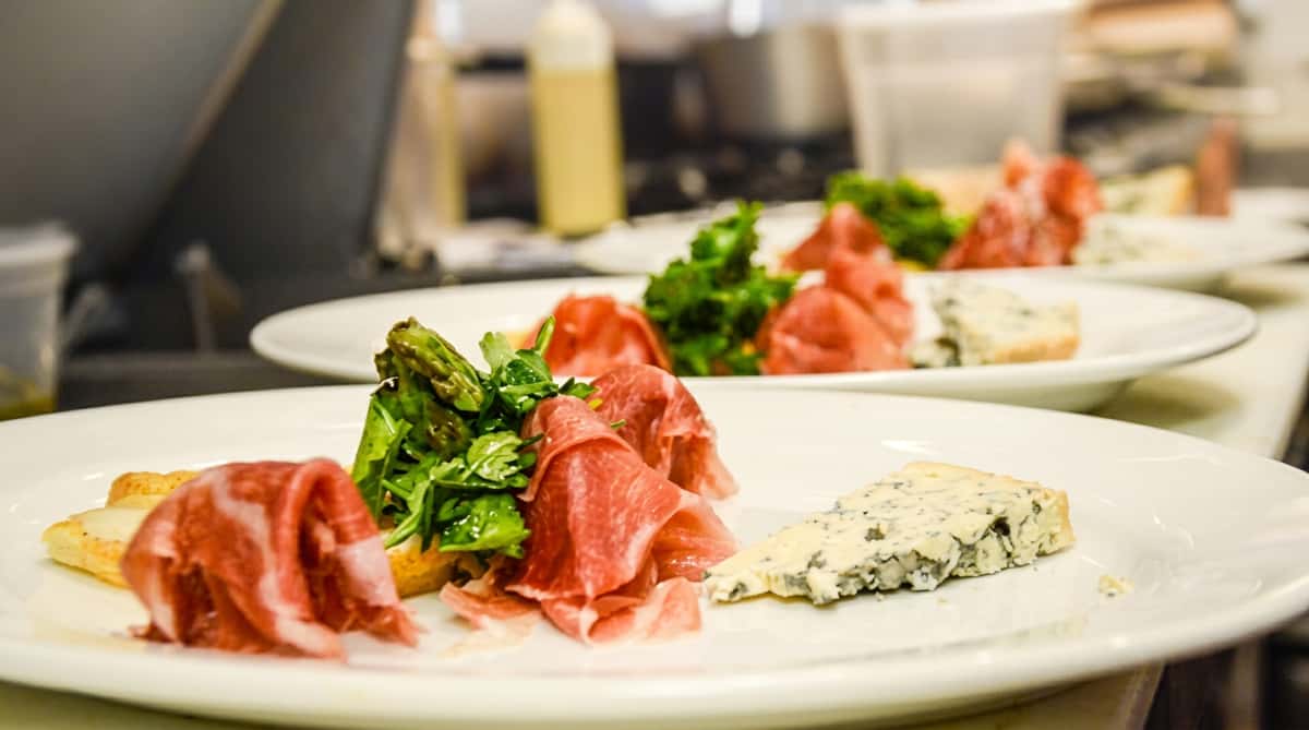 Appetizer plates with prosciutto, greens, and blue cheese on white plates in a kitchen setting.