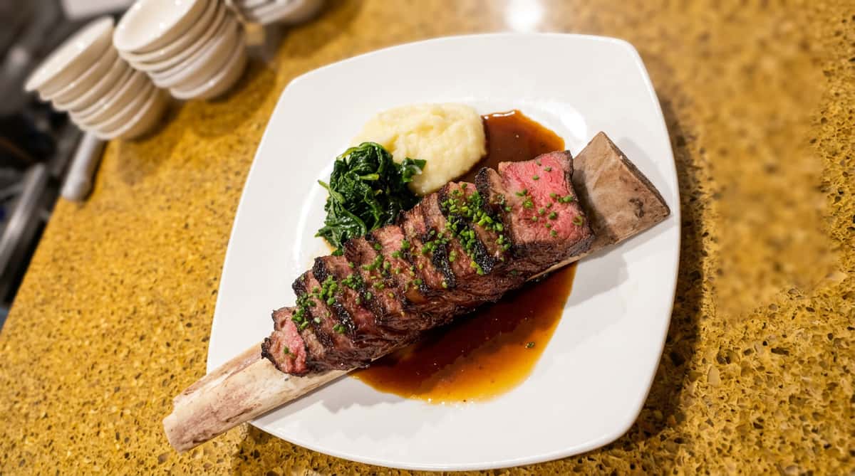 Sliced sirloin steak on bone marrow with mashed potatoes, braised greens, and gravy on a white plate.