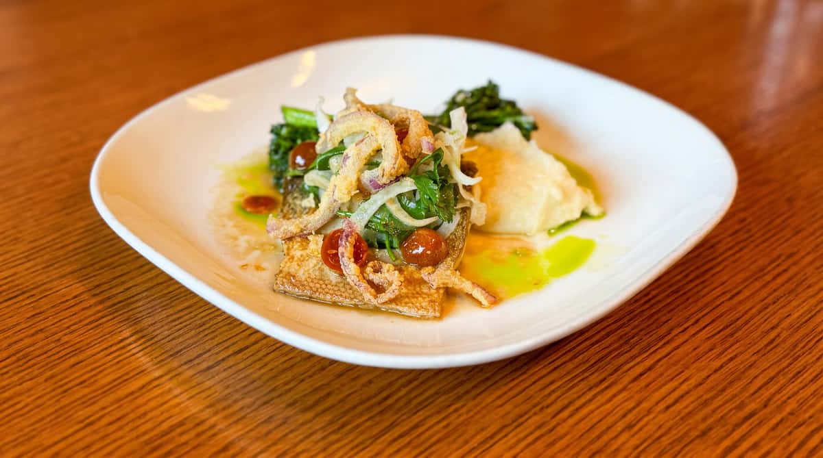 Pan-fried fish fillet with crispy fried onions, colorful salad, and mashed potatoes, drizzled with green oil.
