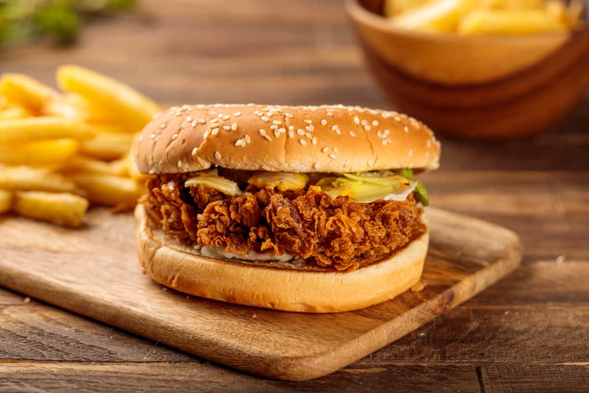 Fried chicken burger on a sesame bun sitting on a wooden board with fries behind it