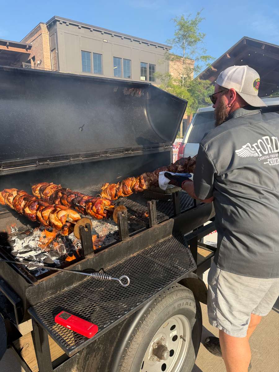 Person tending a rotisserie grill with whole chickens cooking over charcoal.