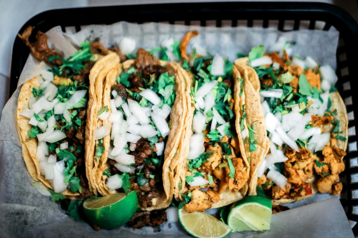 Authentic street tacos served in a black basket, featuring corn tortillas filled with grilled beef and marinated chicken, generously topped with chopped white onions and fresh cilantro. Lime wedges add a zesty finishing touch to this classic Mexican dish.