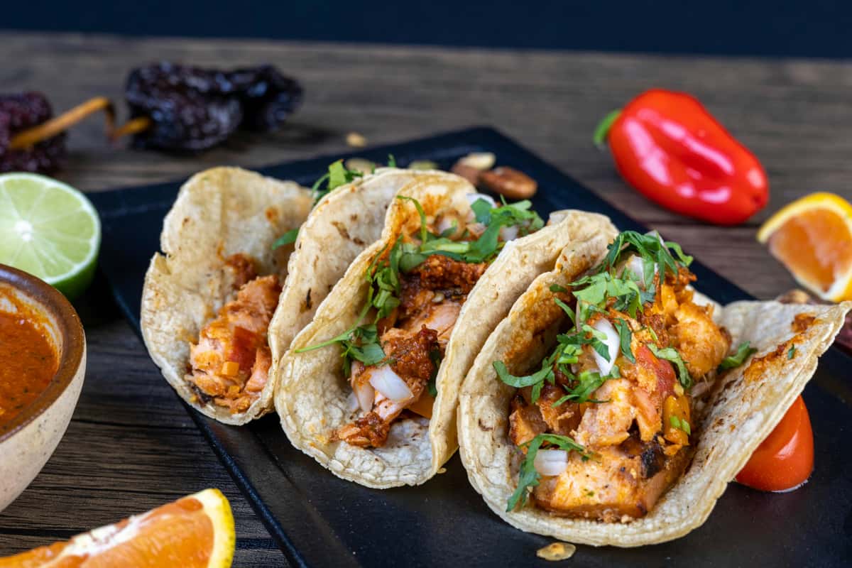 Three grilled fish tacos served on warm corn tortillas, topped with smoky red salsa, chopped onions, and fresh cilantro, plated with citrus wedges and roasted peppers for a bold Mexican street food presentation.