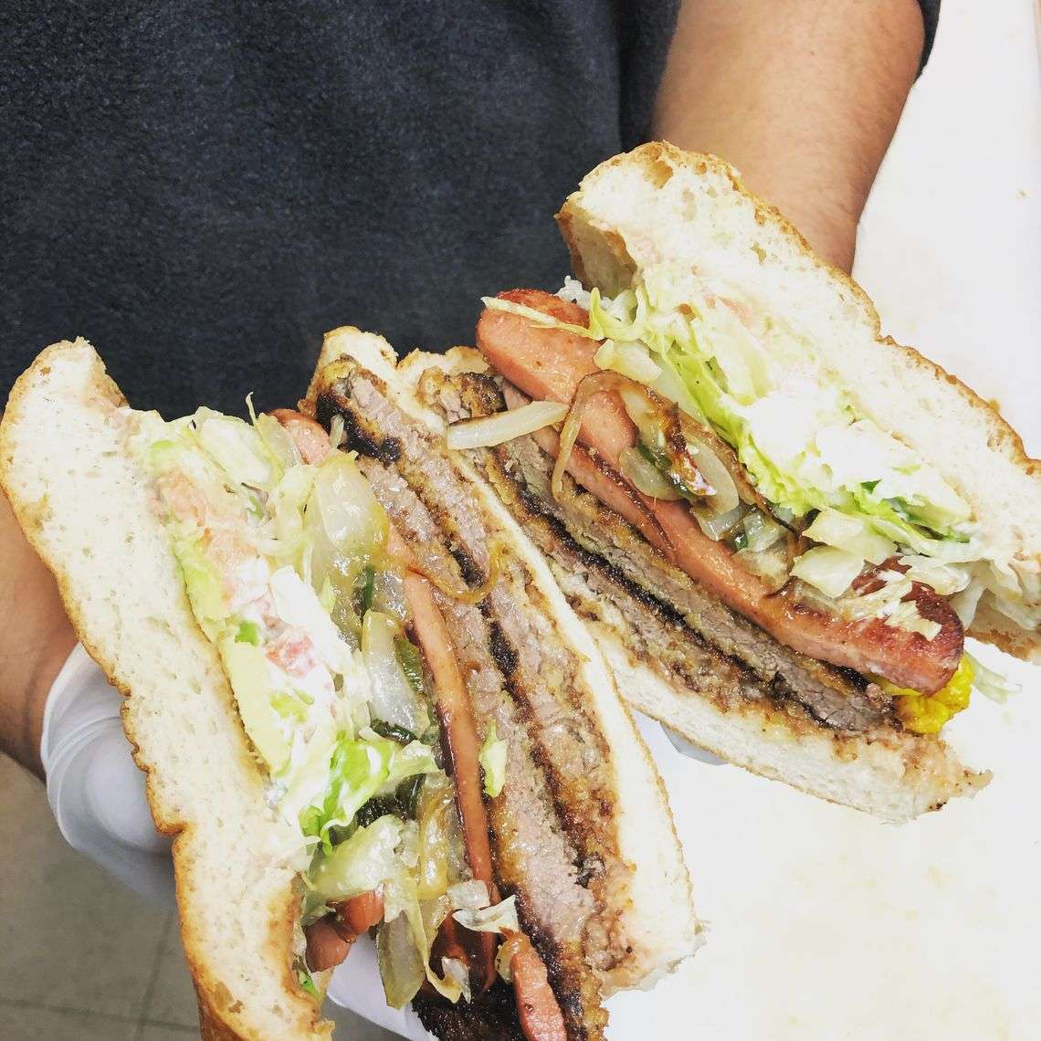 Hearty Mexican torta sandwich sliced in half, loaded with grilled meats, sliced hot dogs, lettuce, tomato, onions, and creamy dressing on soft telera bread—ideal for street food or casual dining menus.