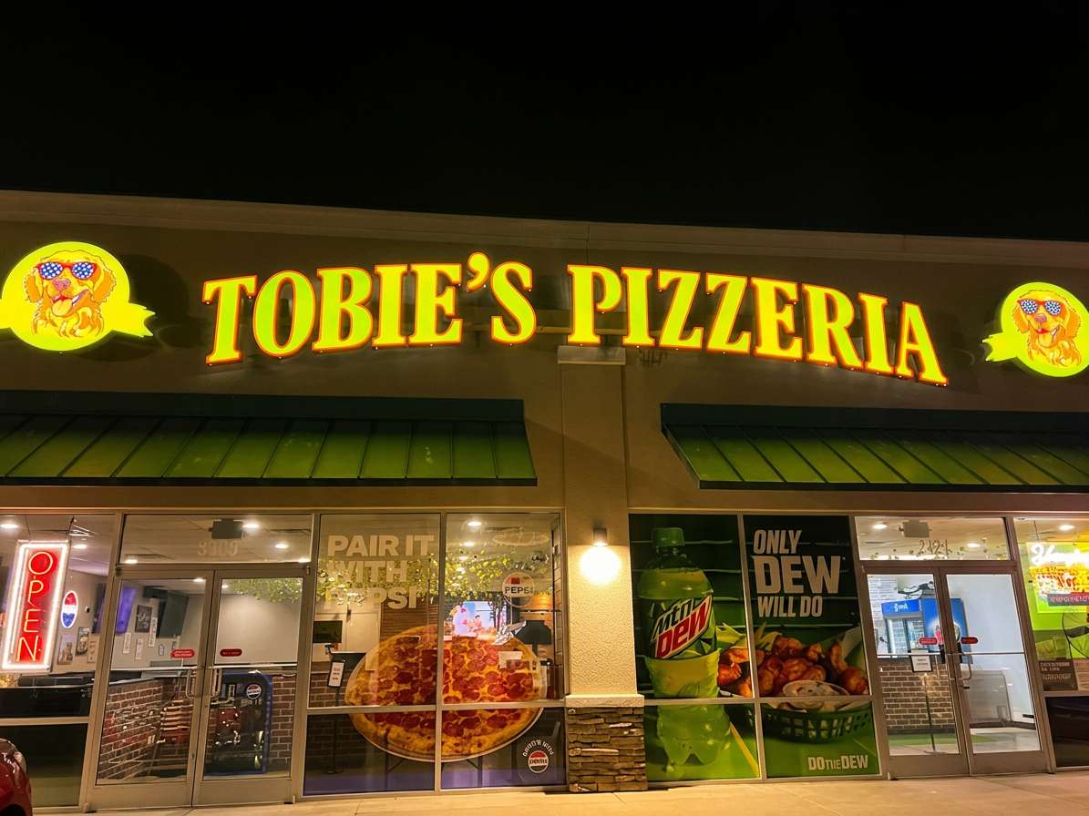 Exterior of Tobie's Pizzeria at night with illuminated storefront signage, welcoming entrance, and family-friendly pizza restaurant featuring arcade and dine-in atmosphere.