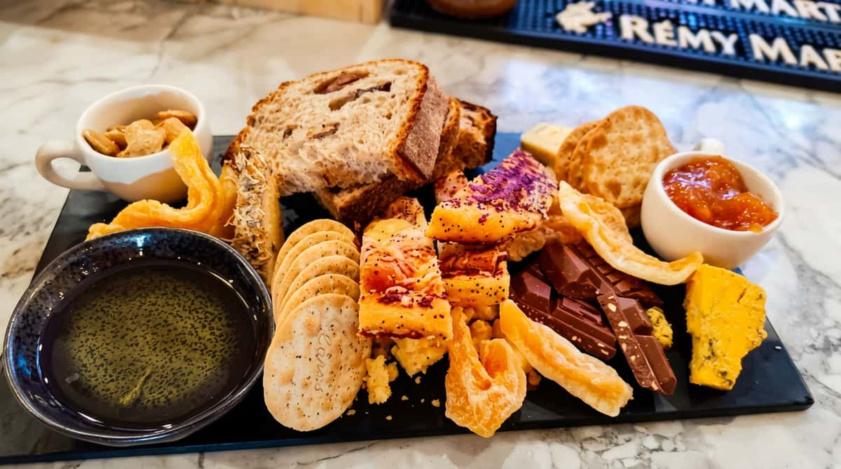 Charcuterie board with sliced bread, various crackers, fruit jam, chocolate, cheeses, and dried fruit.