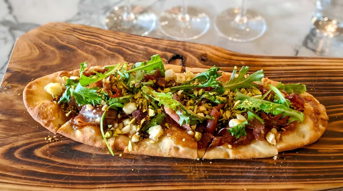 Flatbread with prosciutto, arugula, pistachios, cheese, and balsamic glaze on a wooden board.
