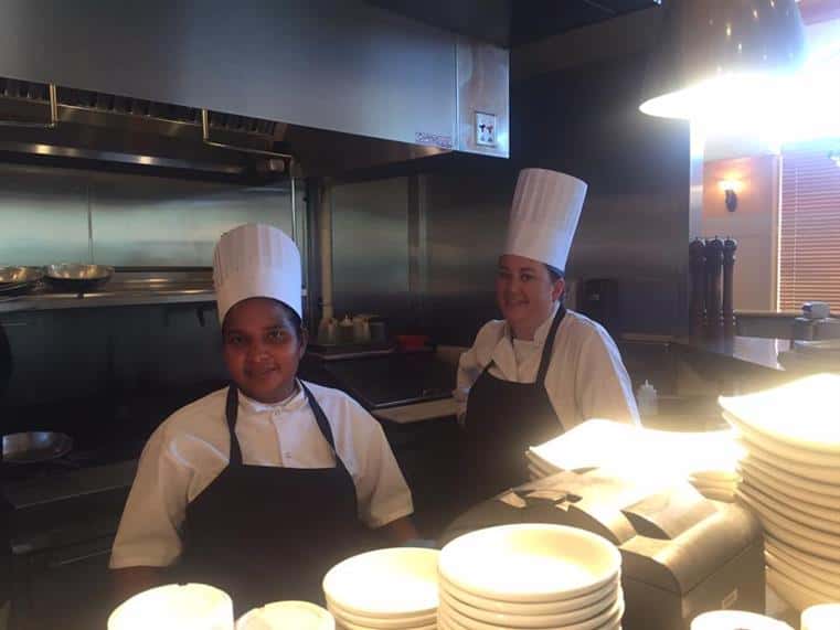 two kitchen staff in kitchen with stacks of plates