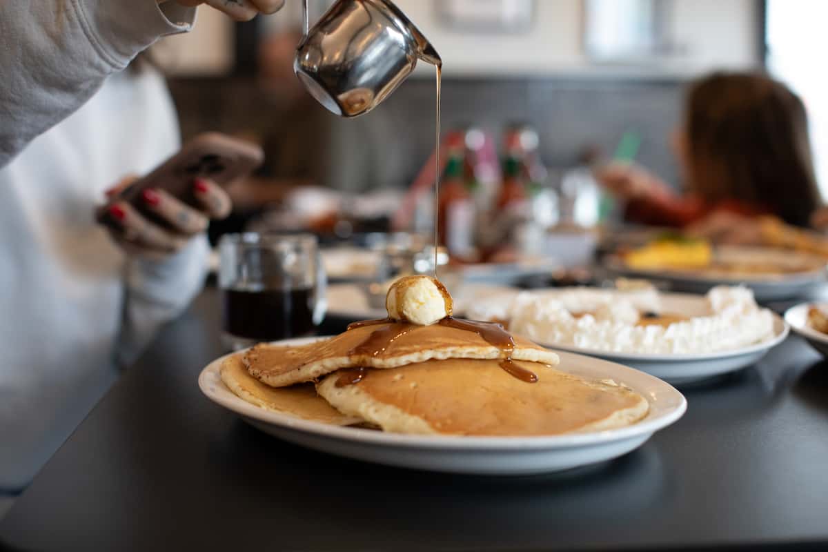 Stack of fluffy golden pancakes topped with a pat of melting butter and drizzled with warm maple syrup, served at a bustling American diner with a cozy, family-friendly atmosphere in the background.