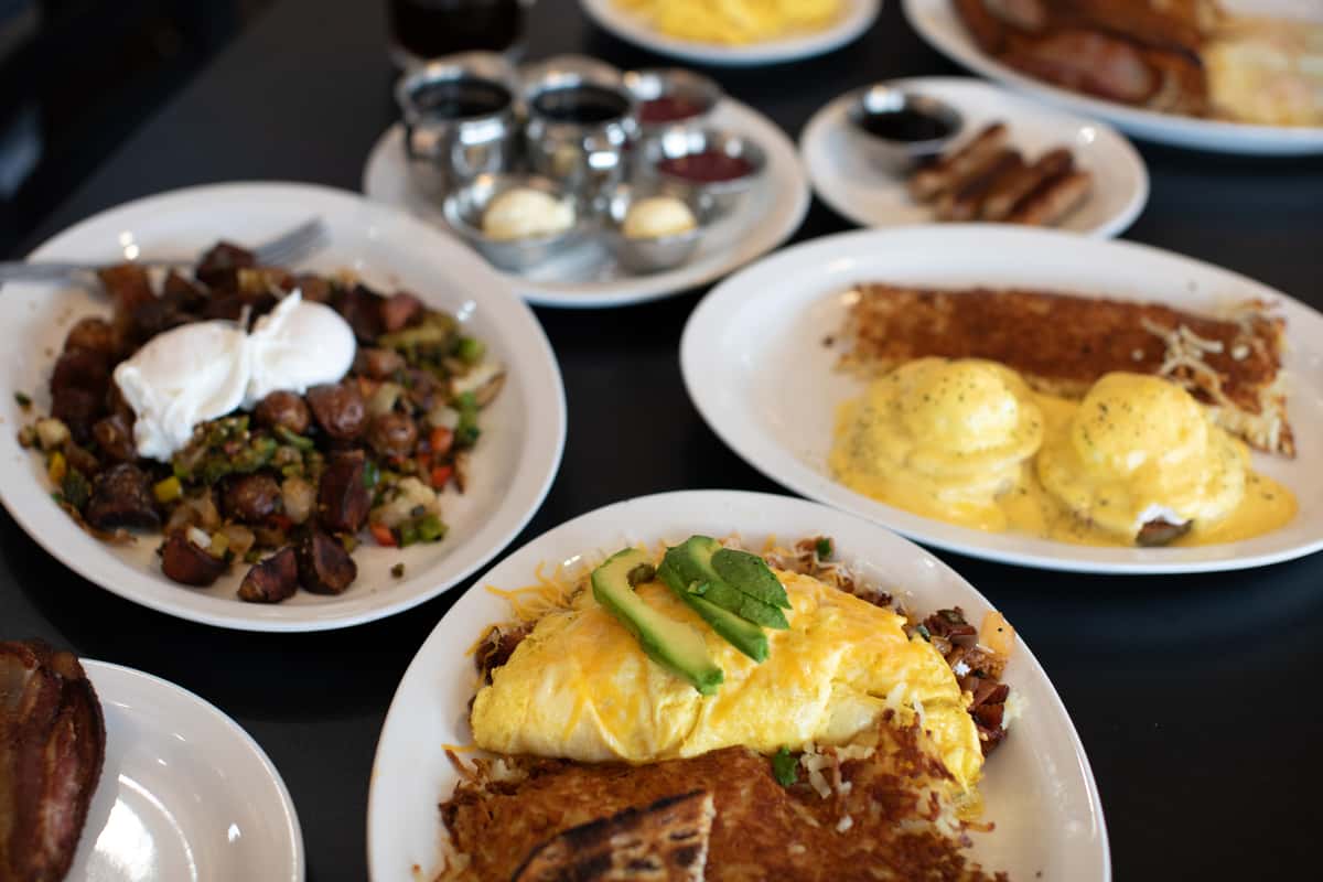 Vibrant American breakfast spread with a fresh avocado-topped omelette, golden hash browns, grilled toast, classic eggs Benedict, seasoned breakfast potatoes with poached eggs, and assorted jams — served at a bustling diner known for hearty, made-from-scratch morning meals.