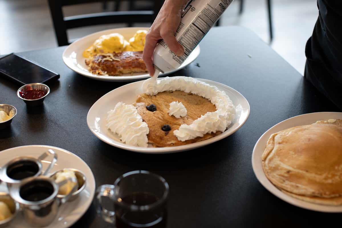 Fun kids' pancake breakfast decorated with whipped cream and blueberries to resemble a smiling face, being finished tableside at a lively American diner — a playful and family-friendly morning meal option.