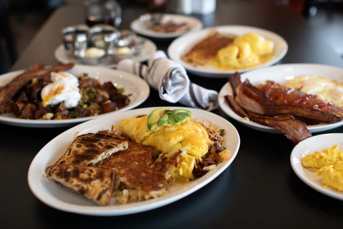Abundant breakfast spread at a classic American diner featuring a fluffy avocado omelette with hash browns and toast, crispy bacon and ham with eggs, a loaded veggie hash topped with poached eggs, and buttery scrambled eggs — perfect for a hearty, made-from-scratch morning feast.