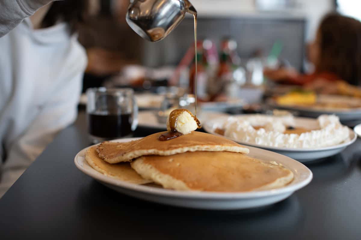 Fluffy stack of buttermilk pancakes topped with a pat of butter and drenched in maple syrup, captured mid-pour at a lively breakfast diner — a comforting and classic morning favorite.