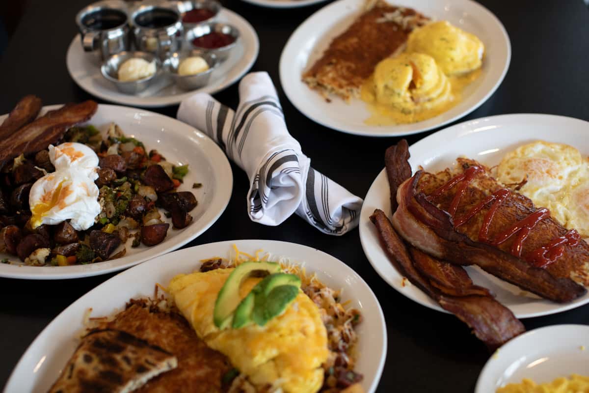 A table filled with breakfast dishes, including eggs Benedict, an omelet topped with avocado, crispy bacon, breakfast potatoes with poached eggs, and pancakes or French toast with syrup, arranged on white plates.