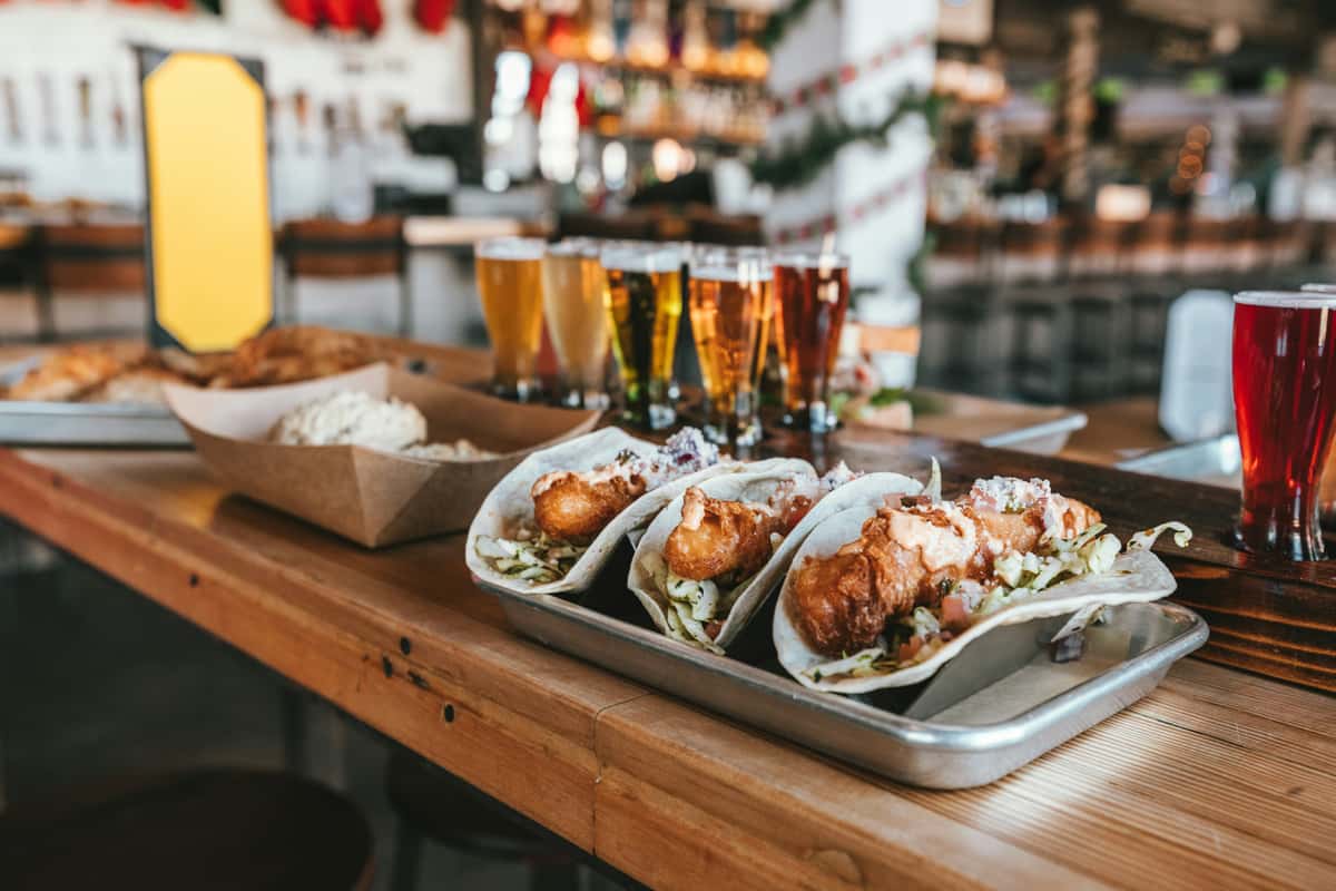 Crispy fish tacos topped with slaw and crema, served with a beer flight of craft brews — fresh Mexican-inspired street food in a modern brewery atmosphere.