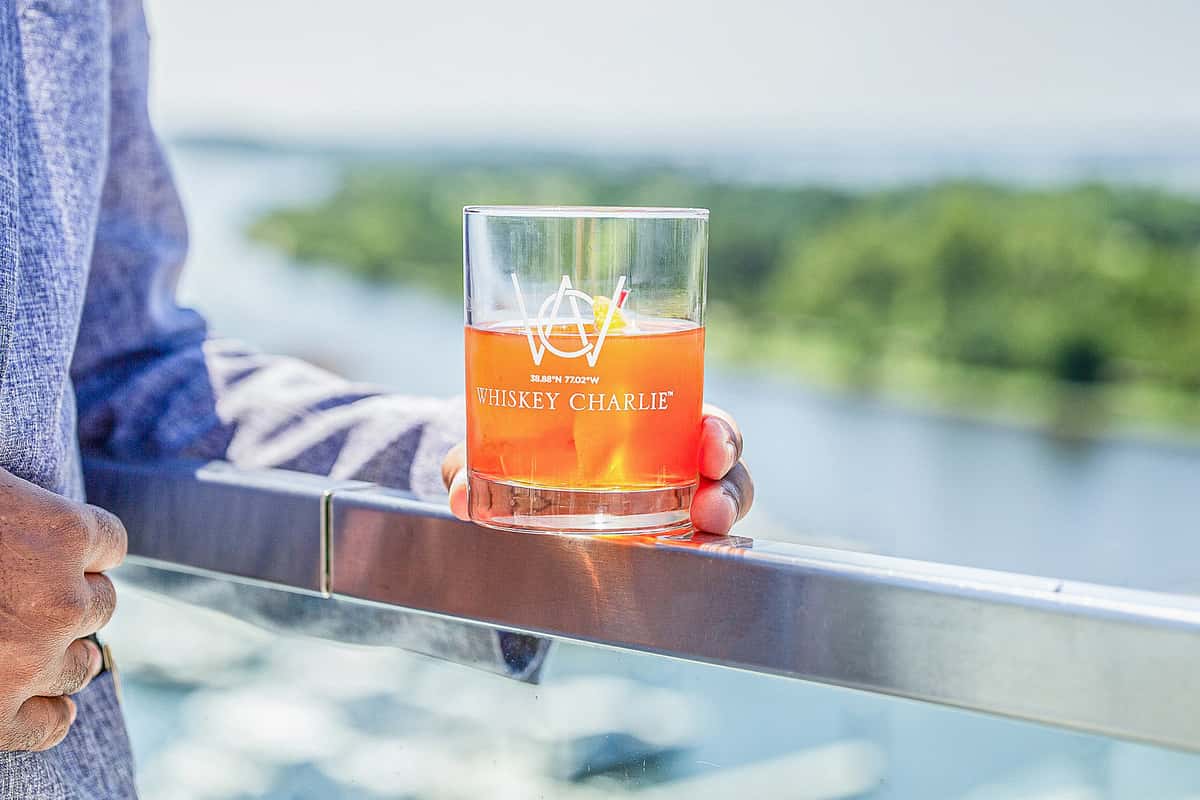 Branded rocks glass with an orange cocktail resting on a metal railing, overlooking a waterfront view at a restaurant