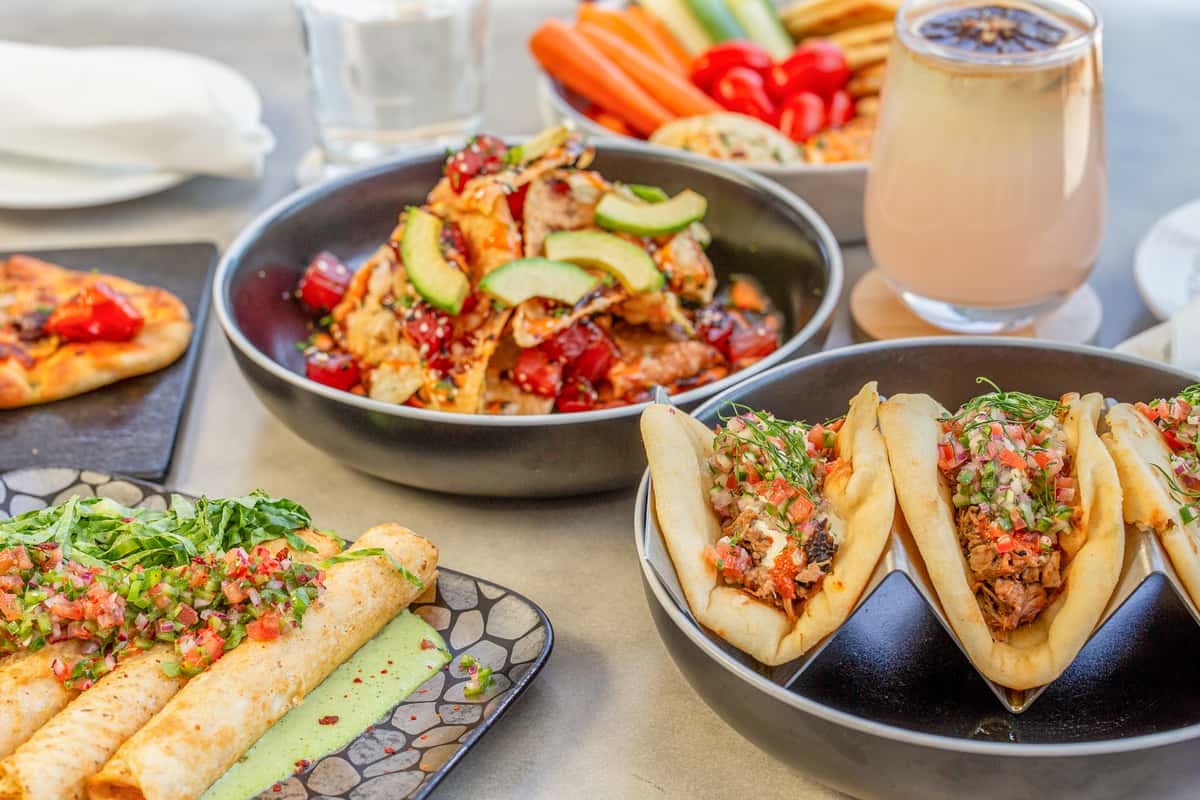 Assortment of restaurant dishes including tacos, nachos, and a wrapped appetizer arranged on a table with a cocktail in the background