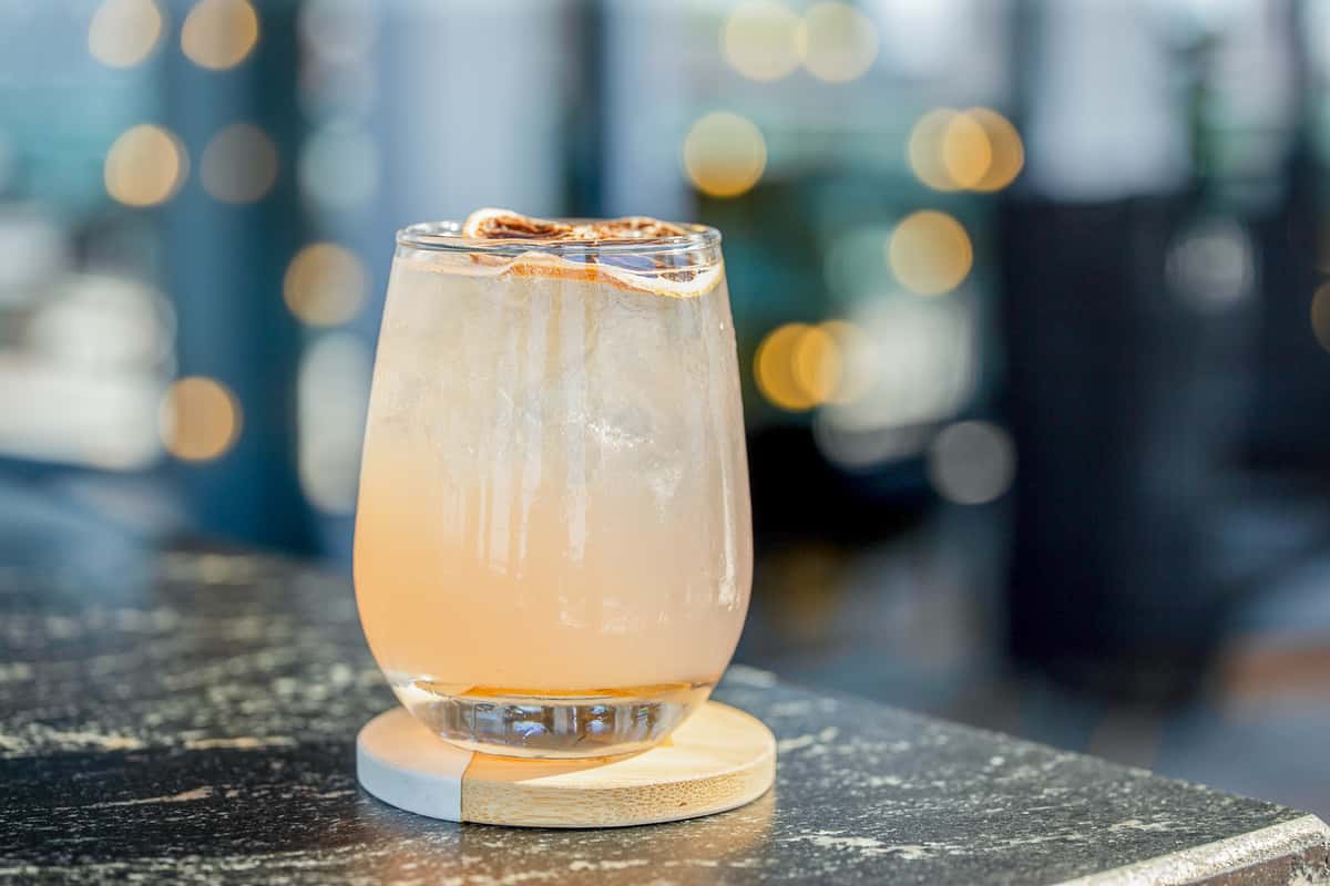 Cocktail with a spiced salt rim served in a rocks glass on a wooden coaster, on a dark table at a restaurant with lights in the background