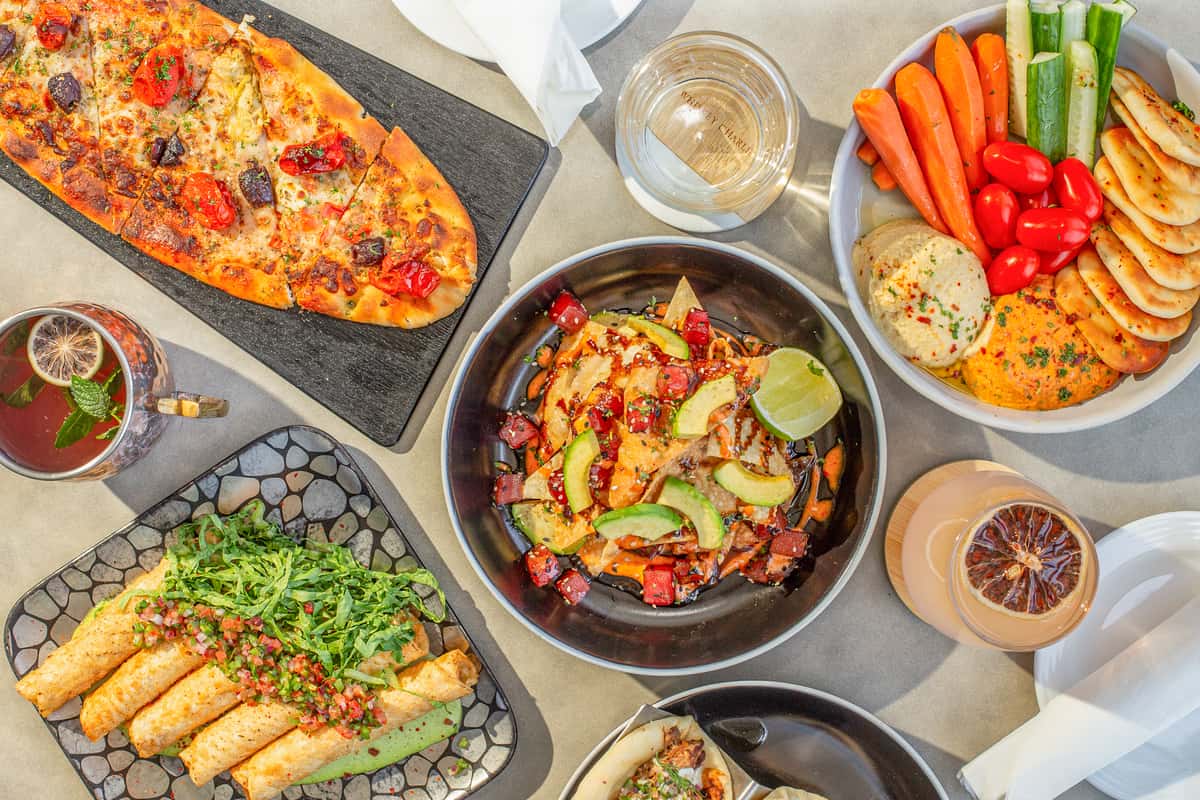 Overhead view of a spread of dishes including flatbread, tuna nachos, taquitos, hummus, and vegetables arranged on a table at a restaurant