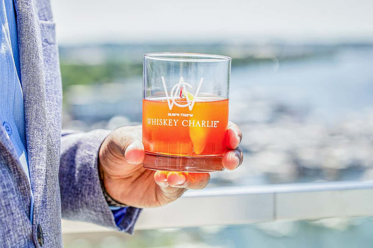 Hand holding a branded rocks glass with an orange cocktail, outdoors with a waterfront view in the background