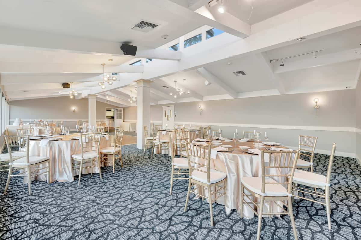 Bright waterfront banquet hall with round tables, gold chiavari chairs, and elegant event setup for weddings and private parties
