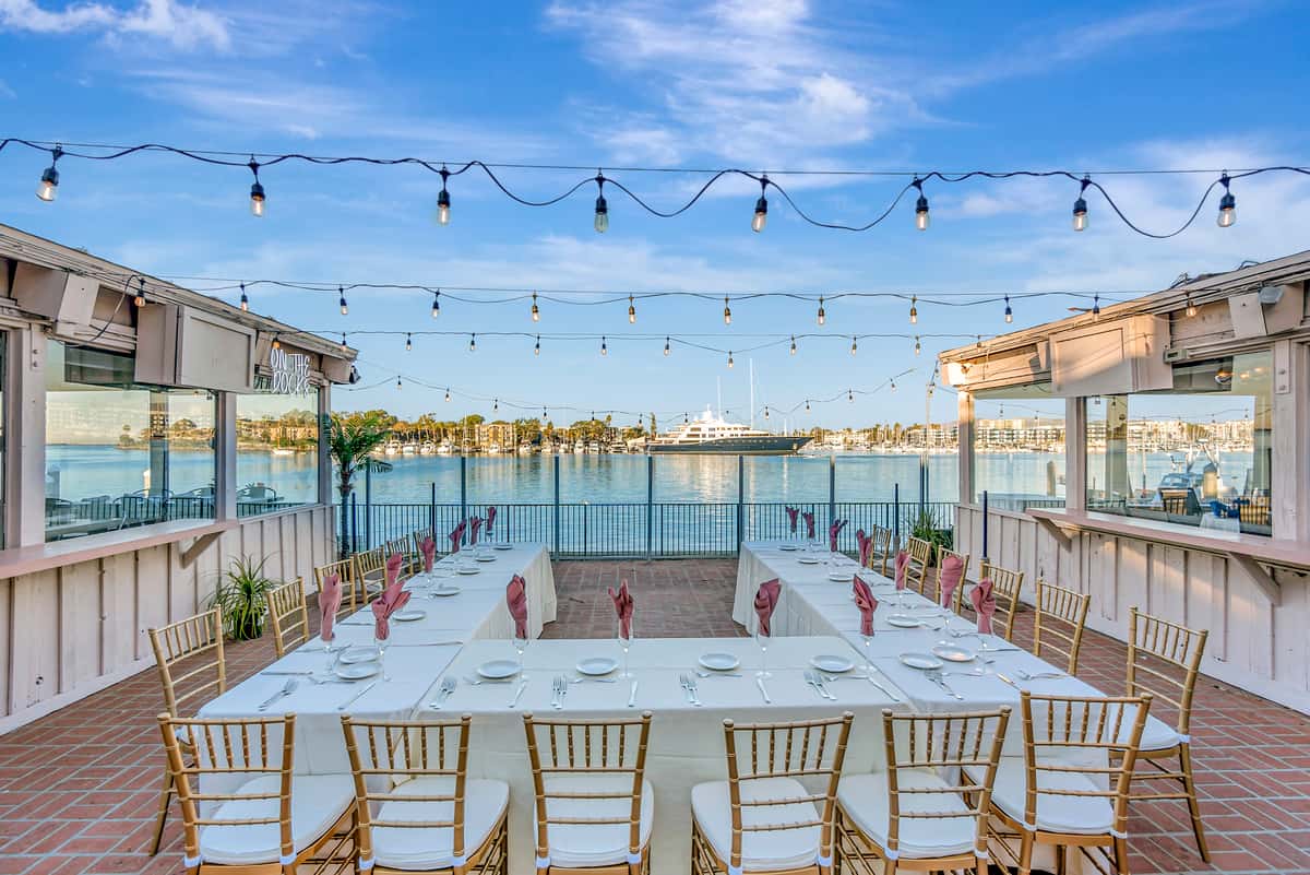 Outdoor waterfront event patio with string lights, banquet seating, and marina views for rehearsal dinners and private parties