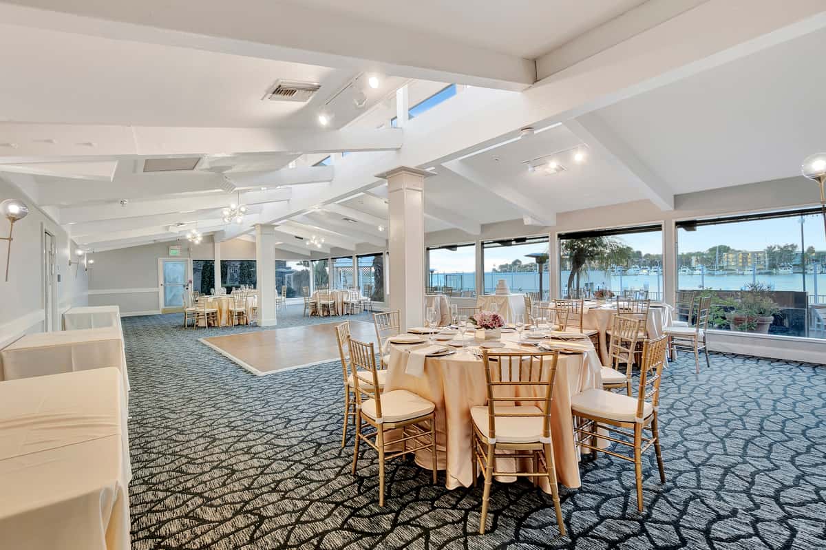 Elegant waterfront ballroom with round banquet tables, gold chiavari chairs, and panoramic bay views for weddings and private events