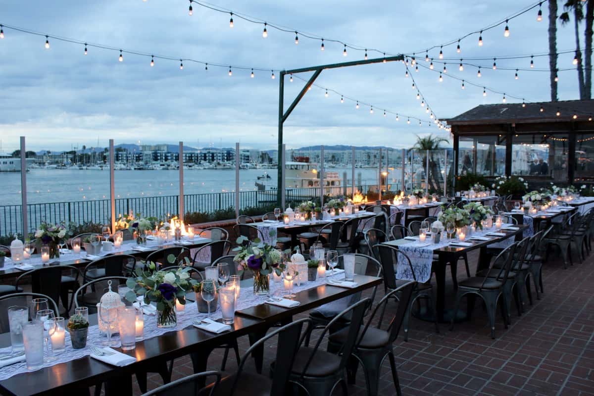 Romantic waterfront reception setup with candlelit banquet tables, string lights, and marina views at an outdoor private event venue