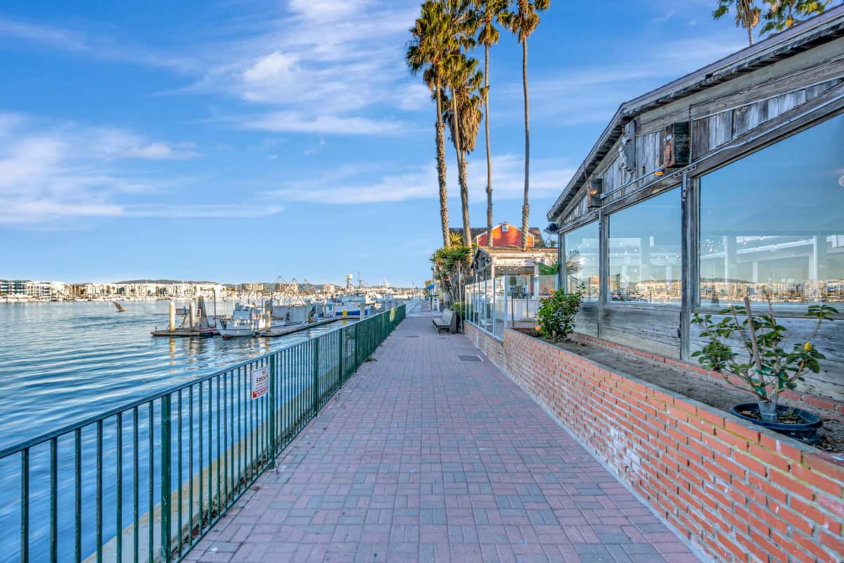 Waterfront boardwalk and marina walkway beside a coastal restaurant with palm trees and harbor views