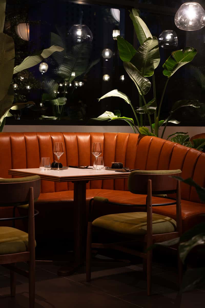 Evening restaurant booth with table settings, pendant lights, and tropical plants