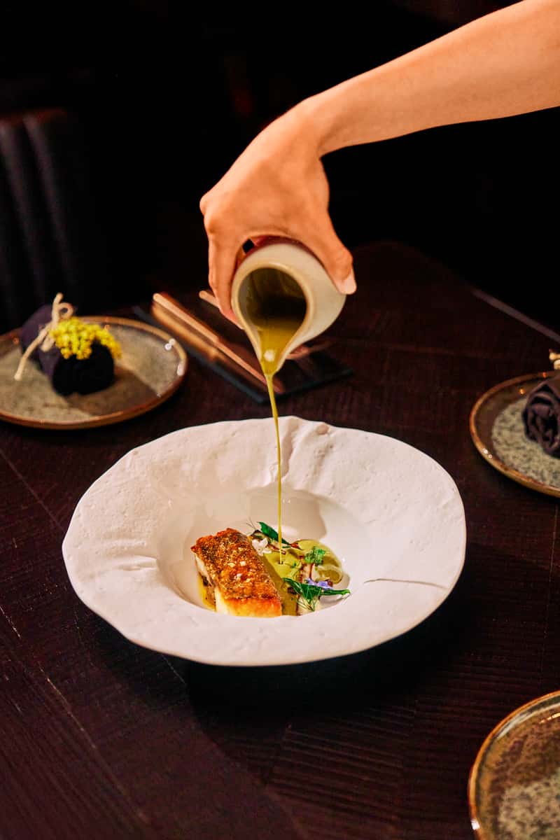 Fine dining fish dish with green sauce being poured tableside