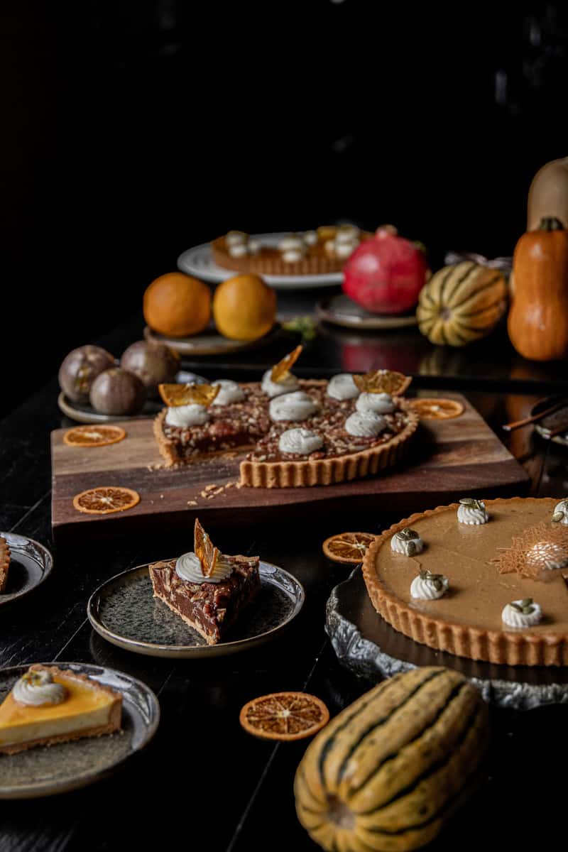Seasonal fall dessert spread featuring pumpkin pie, pecan tart slices, citrus garnishes, and decorative gourds on a dark table