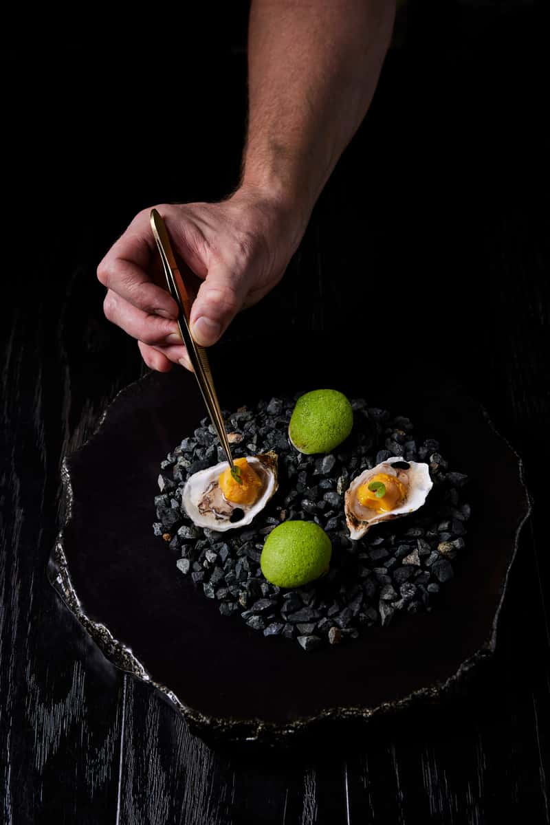 Chef plating oysters with tweezers on a black stone serving plate