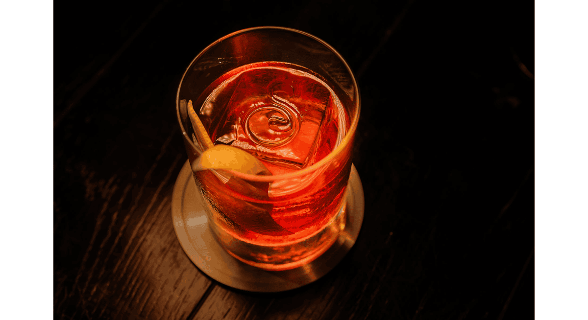 Crafted red cocktail with a large clear ice cube and lemon twist garnish, served in a lowball glass on a coaster in an upscale restaurant setting.