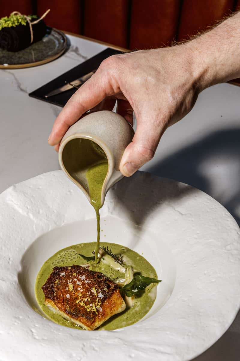 Chef pouring green sauce over a beautifully plated fish dish in a fine-dining restaurant.