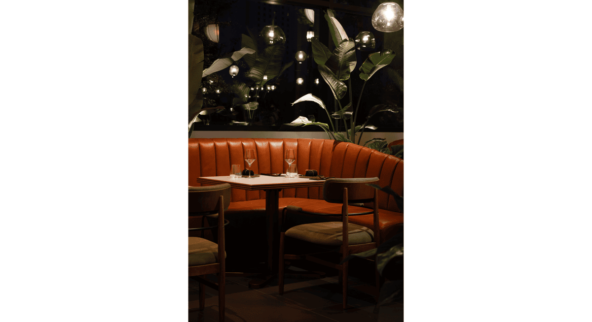 Elegant restaurant booth seating with leather banquette, modern table setting, and indoor tropical plants