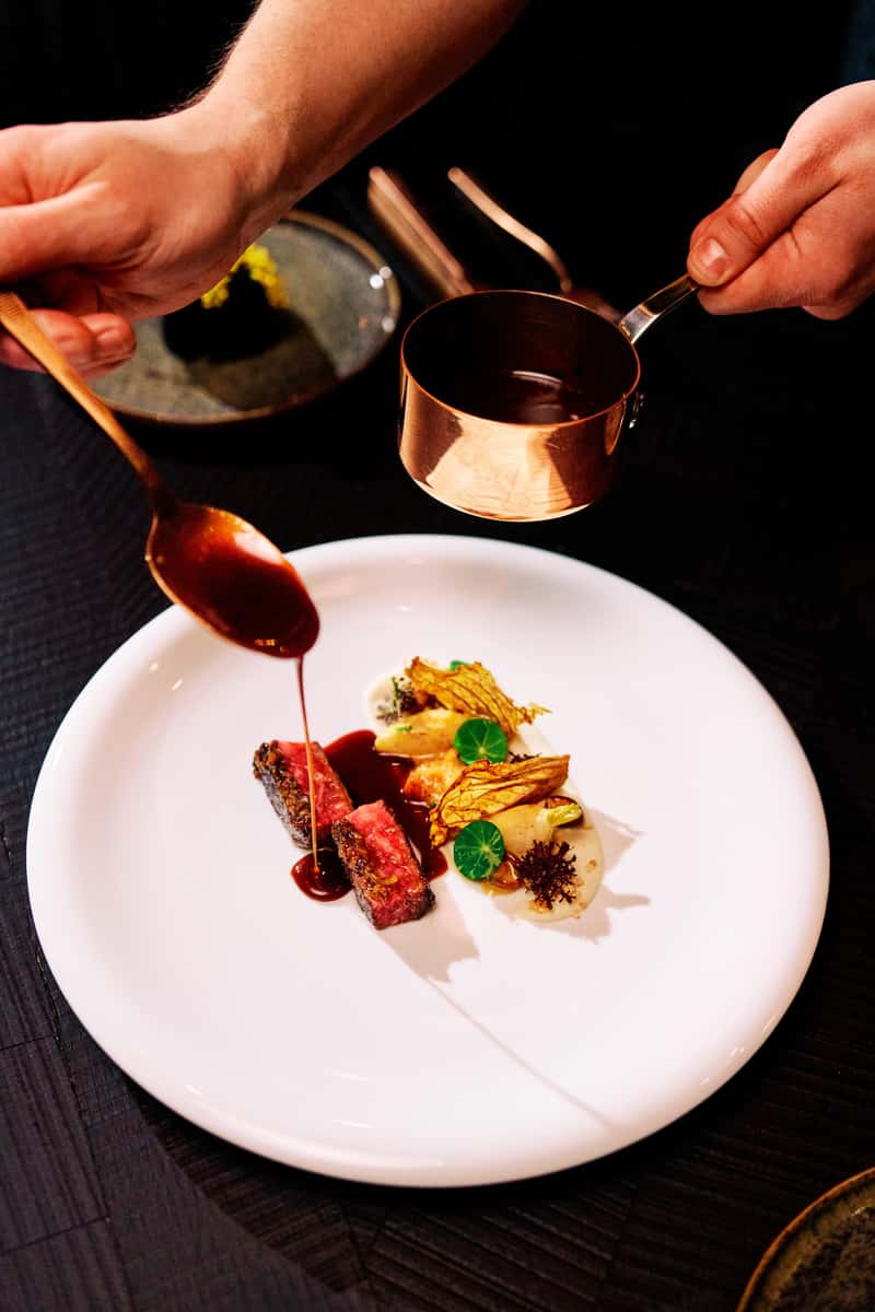 Chef pouring sauce over a gourmet steak dish during fine dining preparation
