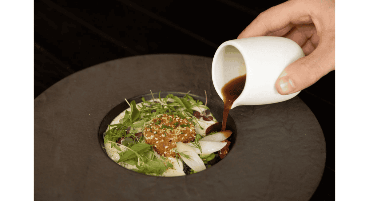 Chef pouring rich sauce over an artfully plated Michelin-star dish with fresh herbs and vegetables at Stubborn Seed Miami.