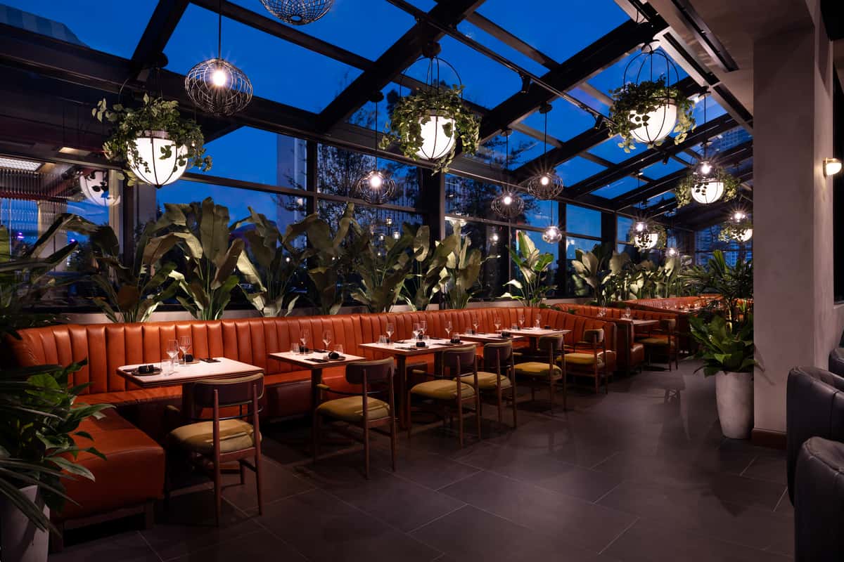 Sophisticated restaurant conservatory with tan leather booths, tropical plants, and hanging lights at dusk.