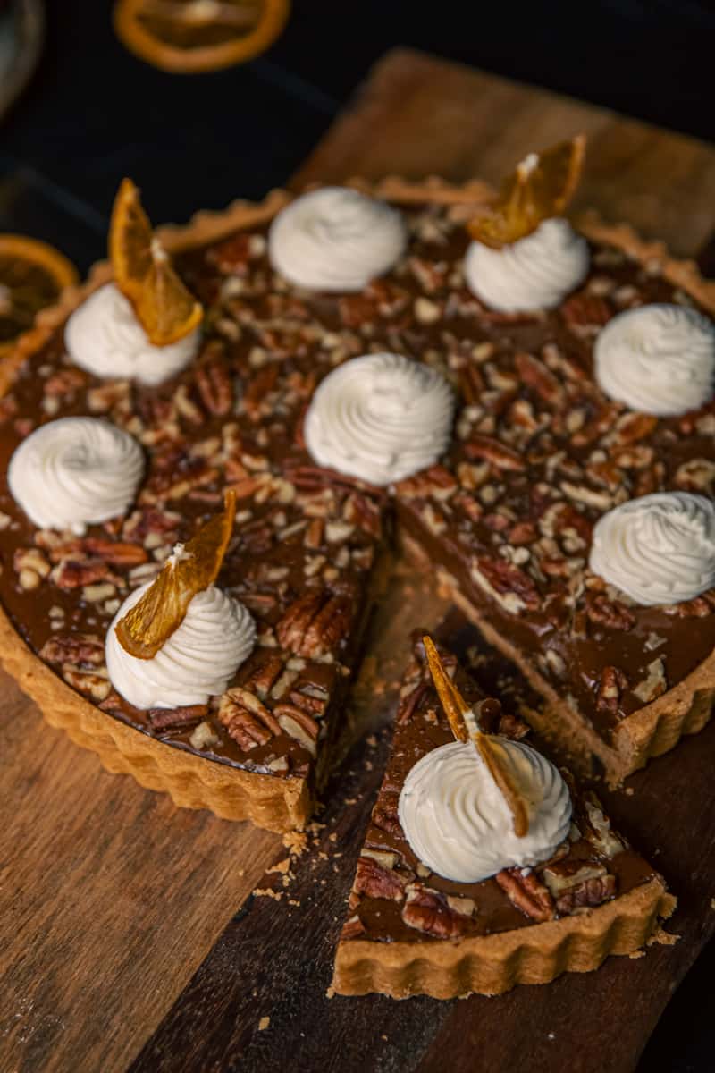 Chocolate pecan caramel tart topped with whipped cream and candied orange slices on a wooden board